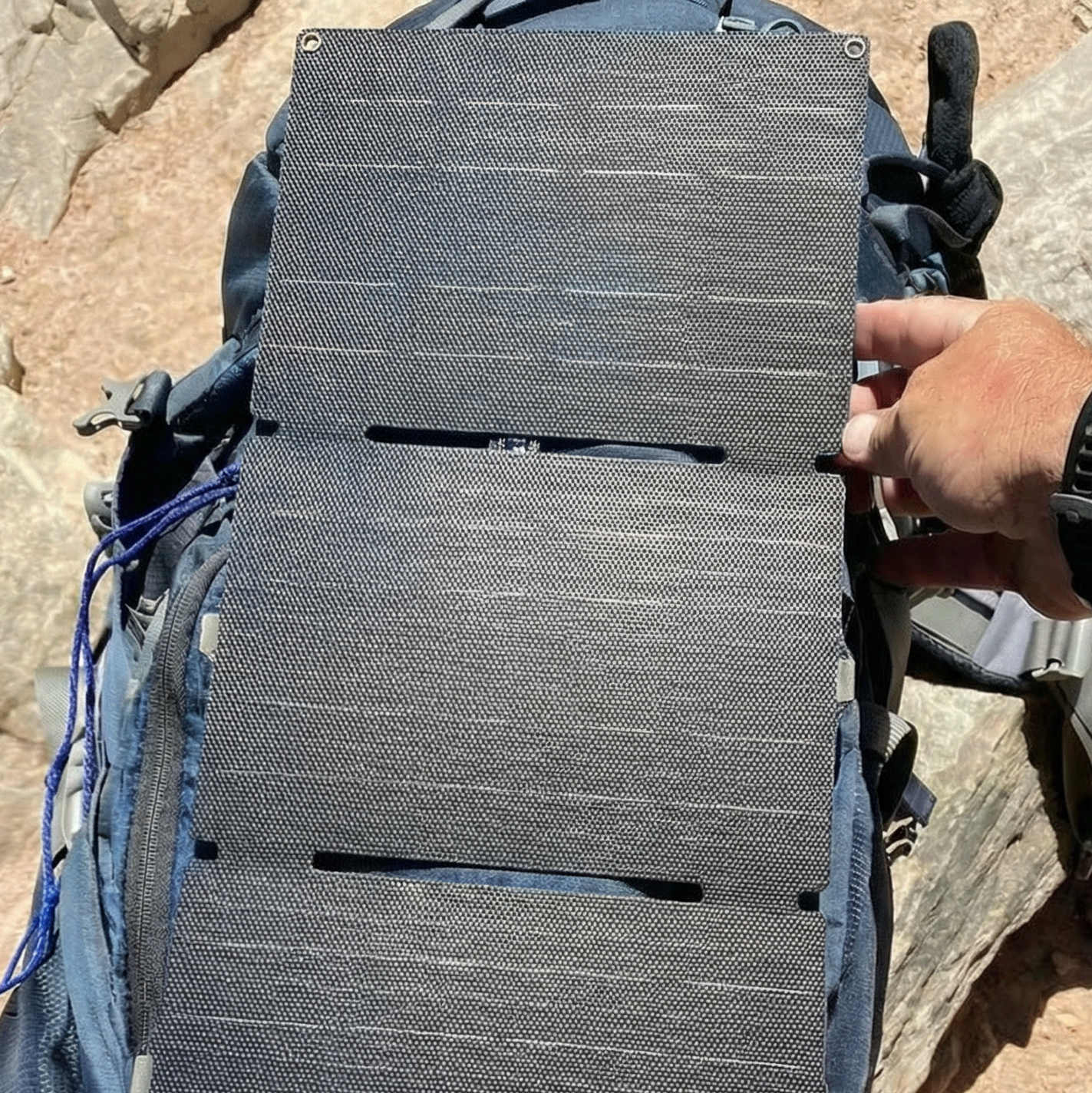 Person holding a solar panel over a backpack on a rocky surface